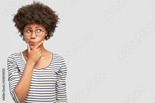 Hesitant thoughtful young African American female holds chin, curves lips, has dark healthy skin, wears striped sweater, looks puzzled aside, contemplates about something, poses over white wall