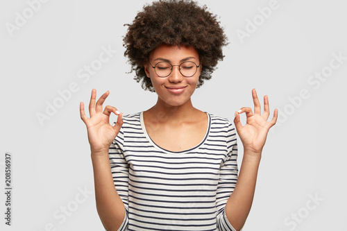 Horizontal shot of happy dark skinned female makes okay gesture, keeps eyes closed from pleasure, demostrates her agreement, has positve expression. Cheerful mixed race young woman poses indoor
