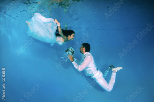 Bride and groom in a white wedding dress swim to each other underwater in the pool. Underwater wedding photo