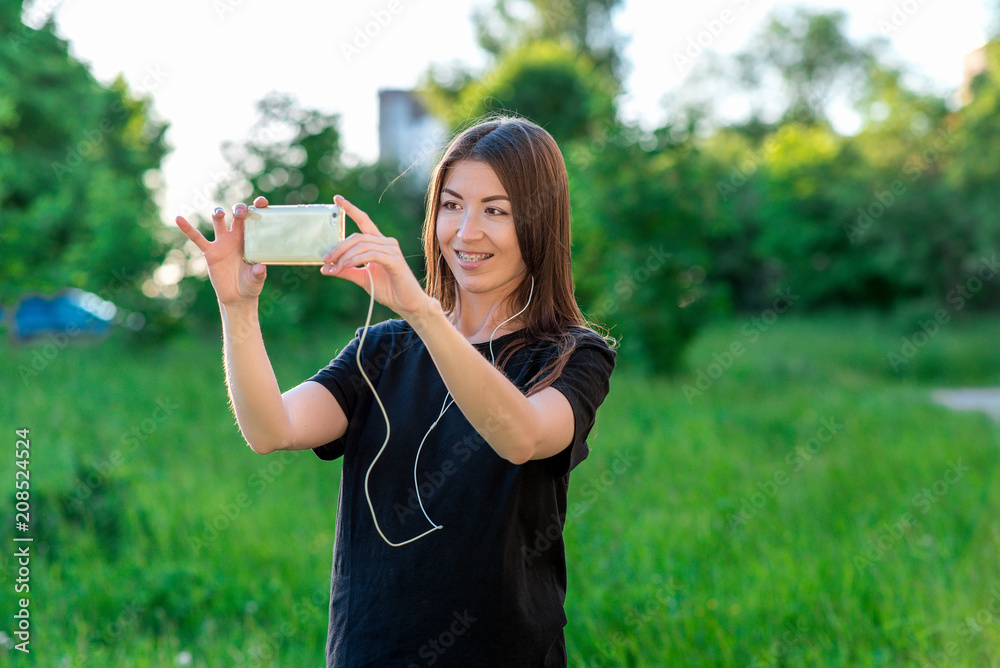 Beautiful happy brunette girl. In summer in park fresh air. Conducts a video broadcast on the social network. Listening to music. Smiles on teeth braces. Photographs in park in the summer. Beautiful happy brunette girl. In summer in park fresh air. Conducts a video broadcast on the social network. Listening to music. Smiles on teeth braces. Photographs in park in the summer.