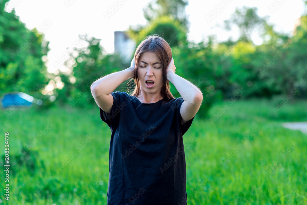 Girl brunette in park outdoors. Hands holding ears. He shouted loudly. Strong loud sound. It is impossible to listen to noise. Emotionally squeezed her eyes slightly open her mouth. Casual clothes. Girl brunette in park outdoors. Hands holding ears. He shouted loudly. Strong loud sound. It is impossible to listen to noise. Emotionally squeezed her eyes slightly open her mouth. Casual clothes.