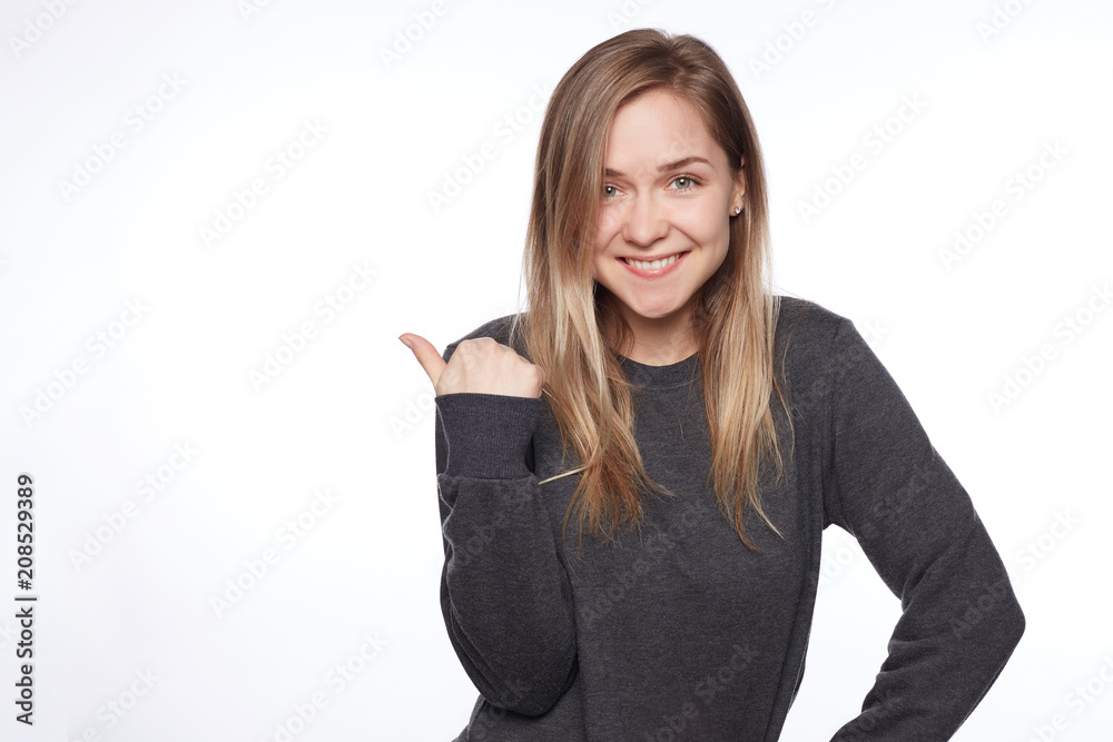 Charming long blond haired teenage lady shows copy space. Pleased lovely woman gives good advice, indicates with thumb, has happy expression. Glad tender female poses indoor over wall. Look there! Charming long blond haired teenage lady shows copy space. Pleased lovely woman gives good advice, indicates with thumb, has happy expression. Glad tender female poses indoor over wall. Look there!