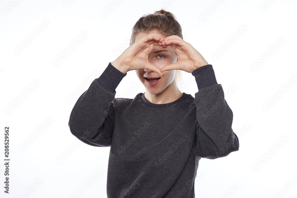 Naklejka premium Young European female with pretty look, blond hair, dressed sporty, shows love sign with hands cupped in heart shape, feels love to someone, stands isolated over white studio wall background.