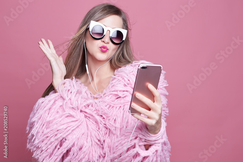 Gorgeous lady dressed in funky bushy coat, wears make up, makes video call via smart phone and headphones, looking at gadget screen, taking selfie sending kiss to boyfriend, standing over pink wall.. photo