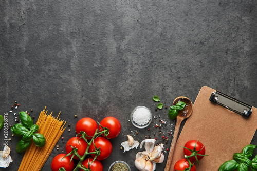 Food background for tasty Italian dishes with tomato. Various cooking ingredients with spoon and blank cardboard clipboard for menu or recipes. Top view with copy space.