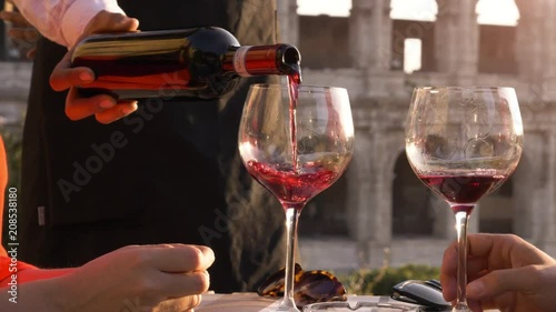Elegant waiter serving pouring glass of red wine to romantic couple sitting at restaurant table in front of colosseum in rome at sunset