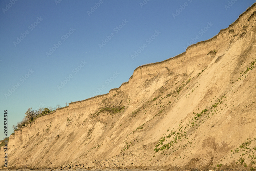Sandy clay soil vertical breakage with plant roots and small isolated vegetation. Blue sky. Sea coast behind the shot Sandy clay soil vertical breakage with plant roots and small isolated vegetation. Blue sky. Sea coast behind the shot