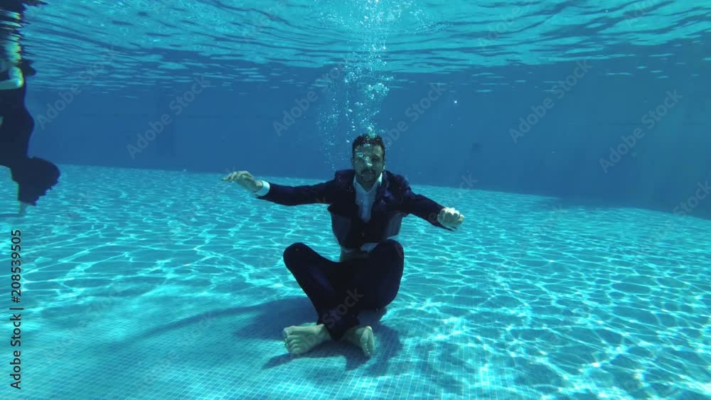 The happy groom in a suit and a white shirt goes down underwater to the bottom of the pool. He sits at the bottom in a Lotus position, looks at the camera and smiles. Slow motion.