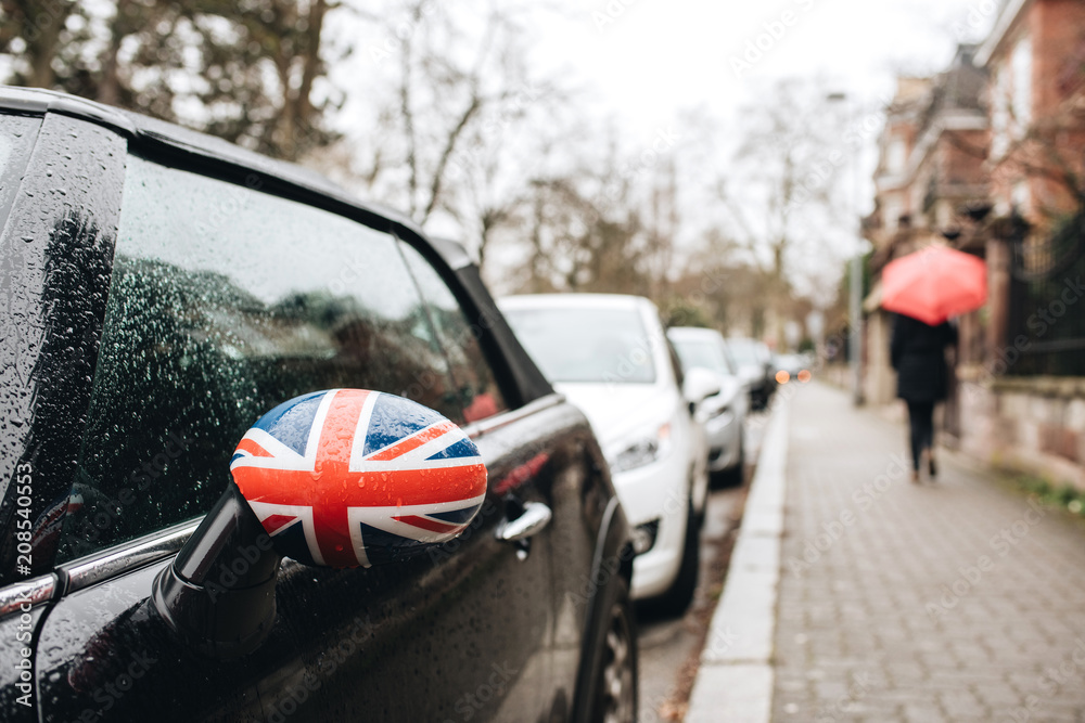 Fototapeta premium British flag Union Jack on a car mirror with silhouette of woman with umbrella on a rainy day in French city walking