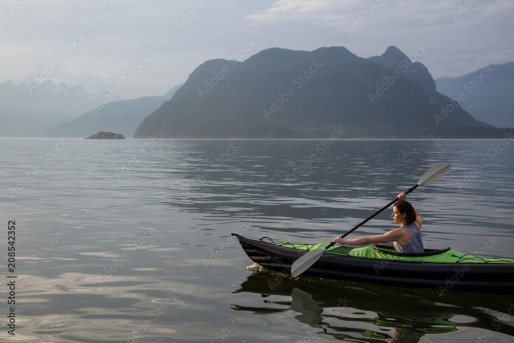 Woman kayaking around the beautiful Canadian Mountain Landscape during a vibrant cloudy evening. Taken in Howe Sound, North of Vancouver, British Columbia, Canada.