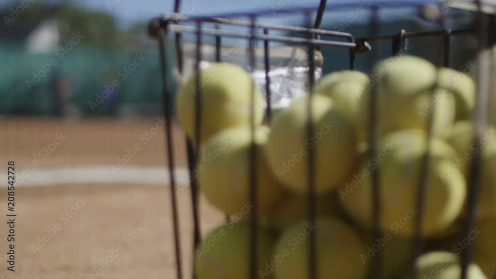 Metal bucket with yellow fluffy tennis balls on sports ground of tennis court in bright sunlight