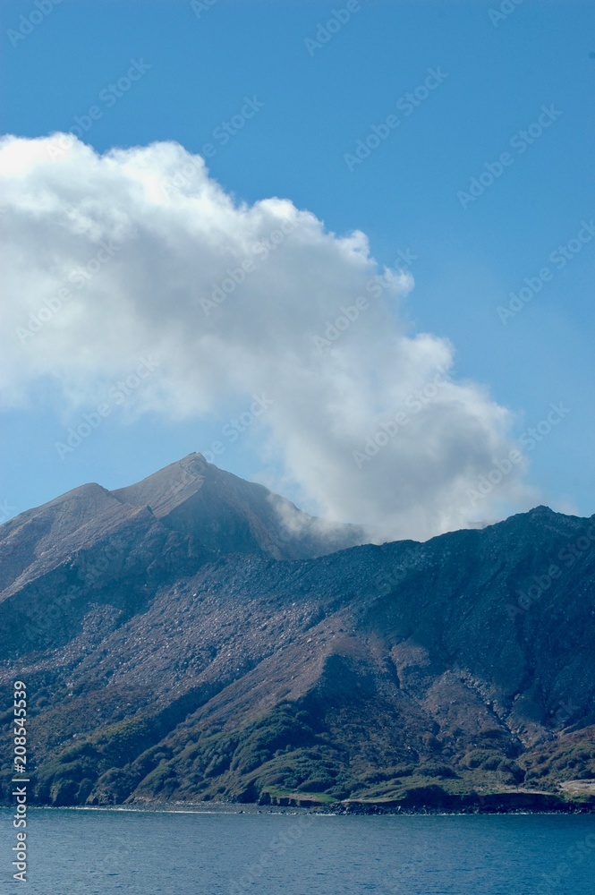 An active volcano, seen from the sea. Smoke is rising into a clear blue sky.