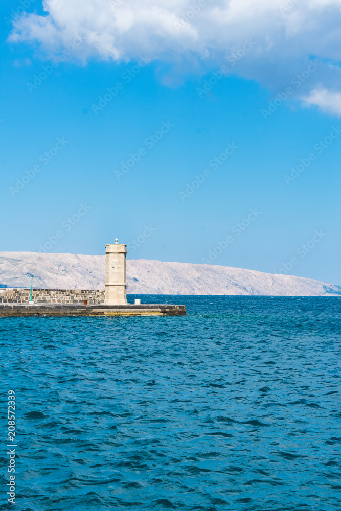 Seascape view of an old stone lighthouse at the end of a stone pear with colorful green lantern in contrast with the clear blue Adriatic sea in Croatia, town of Senj. Summer in Croatia concept Seascape view of an old stone lighthouse at the end of a stone pear with colorful green lantern in contrast with the clear blue Adriatic sea in Croatia, town of Senj. Summer in Croatia concept