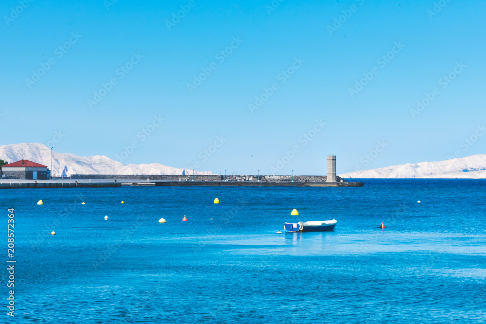 View of a small fisherman's boat in front of a stone pear with an old lighthouse with white islands in the background. Blue Adriatic sea in Croatia. Summer in Croatia or travel concept
