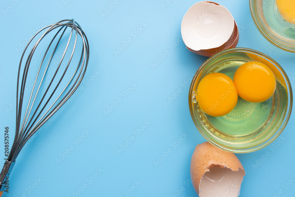 Fototapeta premium Raw fresh egg in glass bowl with eggshell and whisk on blue background top view with copy space