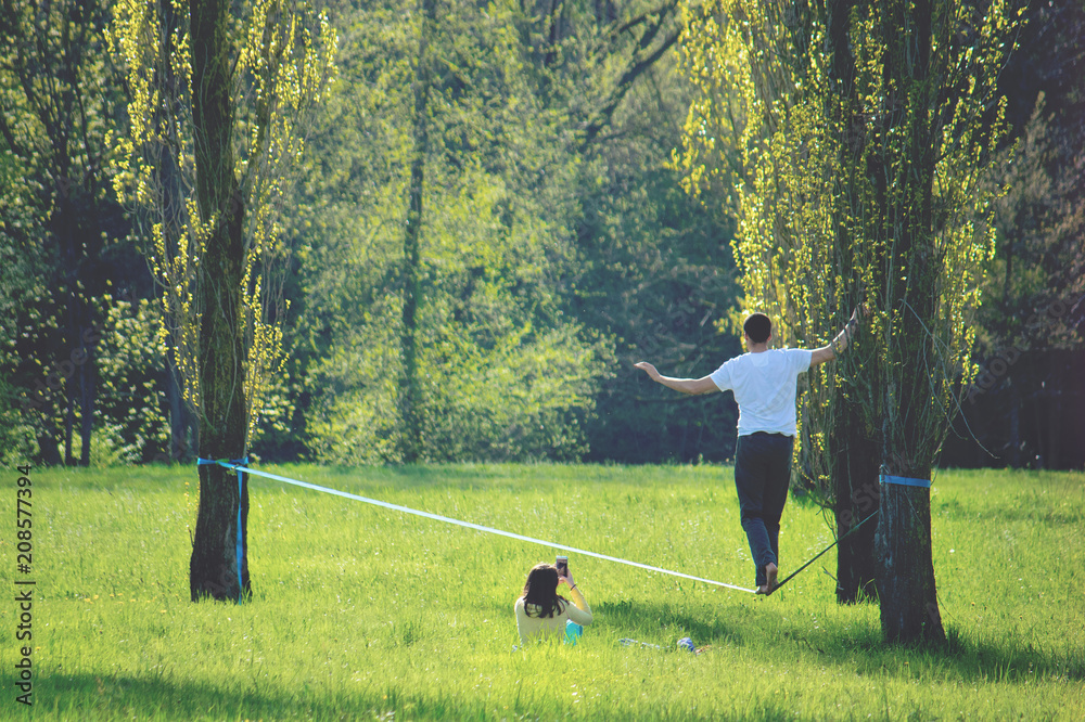 young man walking on a wire, a girl sitting on the green grass shooting the slackling activity with her smartphone
