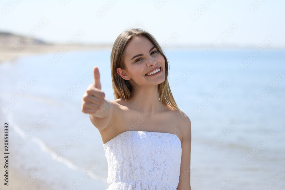 Hübsche blonde Frau steht lachend an einem Strand und zeigt Daumen hoch Hübsche blonde Frau steht lachend an einem Strand und zeigt Daumen hoch