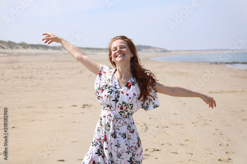 Hübsche rothaarige Frau steht mit ausgebreiteten Armen und geschlossenen Augen lächelt an einem Strand