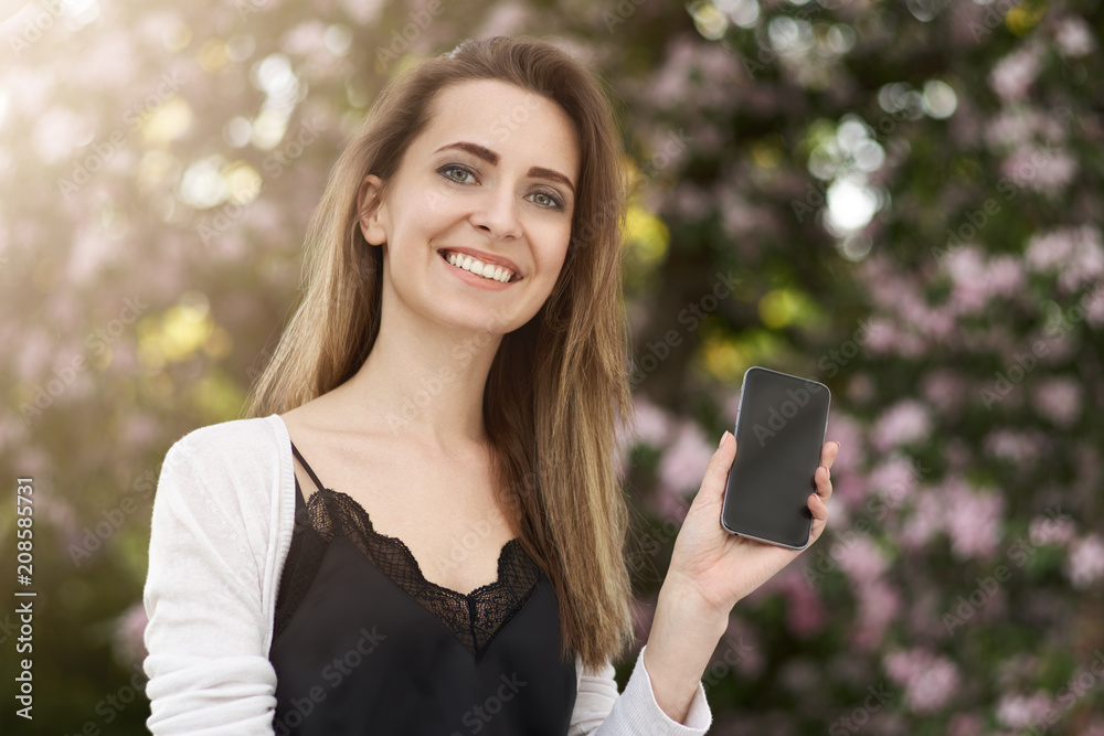 smiling long-haired girl on the background of green foliage of trees holding a smartphone with an empty display smiling long-haired girl on the background of green foliage of trees holding a smartphone with an empty display