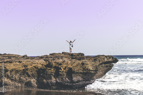 Young woman on the rock on a black sand beach, Bali island. Freedom concept.