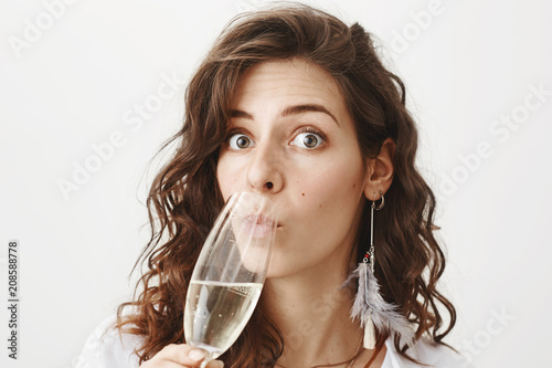 Close-up portrait of cute surprised caucasian female drinking champagne from glass and glaring at camera with intrigued expression, being caught with alcohol, standing over gray background