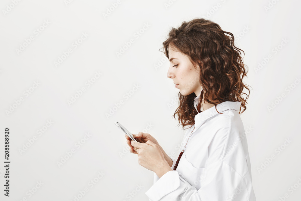 Profile picture of sensual caucasian woman with fashionable hairstyle holding smartphone while looking at it with serious expression, being focused and standing over gray background. Profile picture of sensual caucasian woman with fashionable hairstyle holding smartphone while looking at it with serious expression, being focused and standing over gray background.