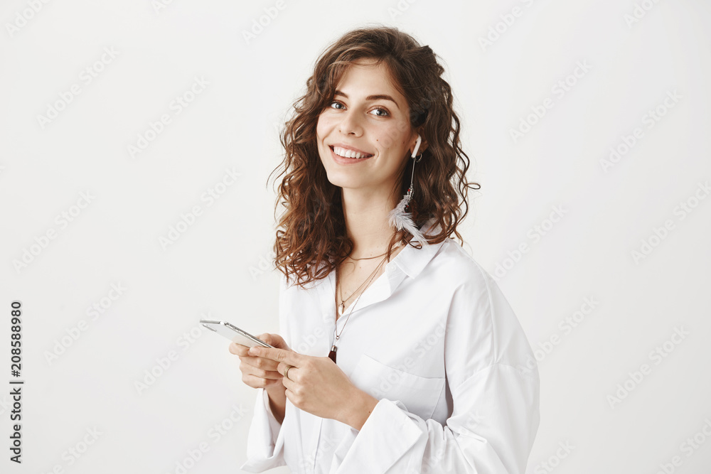 Portrait of gorgeous adult successful woman in white blouse holding smartphone while smiling broadly at camera, standing over gray background and listening music in wireless earphone. Portrait of gorgeous adult successful woman in white blouse holding smartphone while smiling broadly at camera, standing over gray background and listening music in wireless earphone.