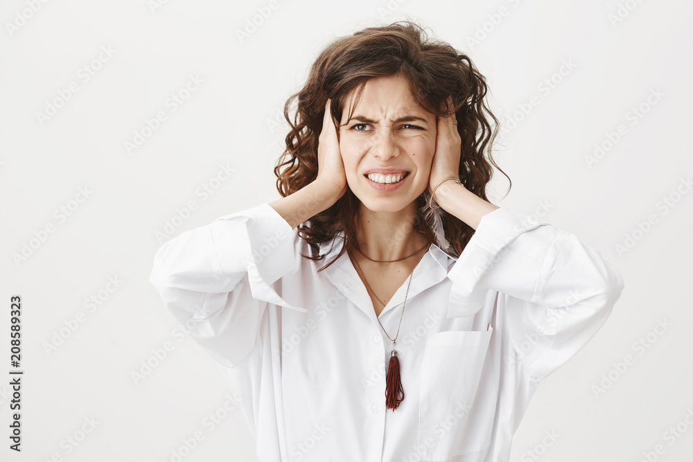Studio portrait of annoyed unsatisfied attractive caucasian female covering ears with hands, looking aside and frowning while standing over gray background. Girl distracted by irritating sound