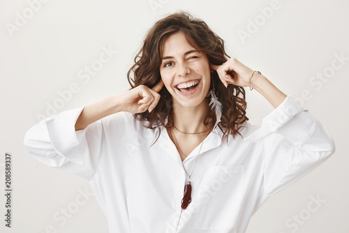 Studio portrait of playful emotive cute adult woman covering ears with index fingers while winking and smiling broadly at camera, standing against gray background. Say what you want I am not listening