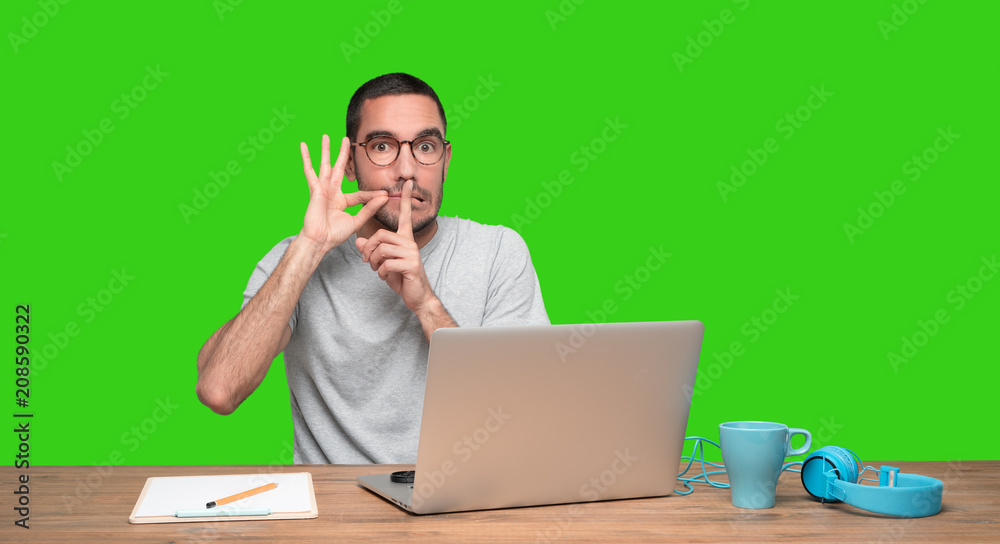 Confident young man sitting at his desk with a gesture of keeping a secret Confident young man sitting at his desk with a gesture of keeping a secret