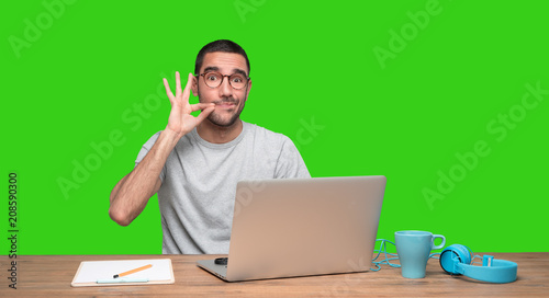 Confident young man sitting at his desk with a gesture of keeping a secret photo