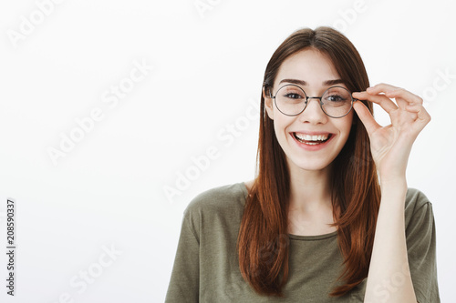 Girl ready to start working on new chapter of book. Indoor shot of creative attractive young woman in trendy round glasses, holding rim and smiling broadly, standing over gray background