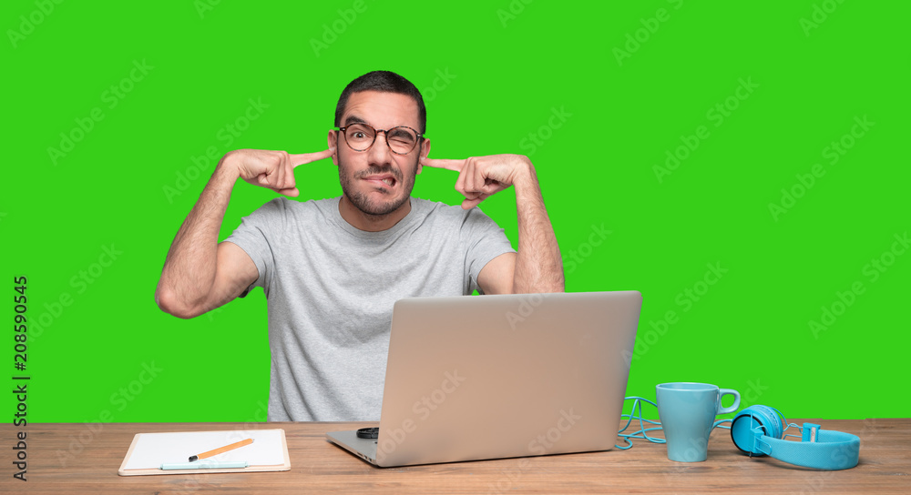 Worried young man sitting at his desk and covering his ears with his hands - Green background Worried young man sitting at his desk and covering his ears with his hands - Green background