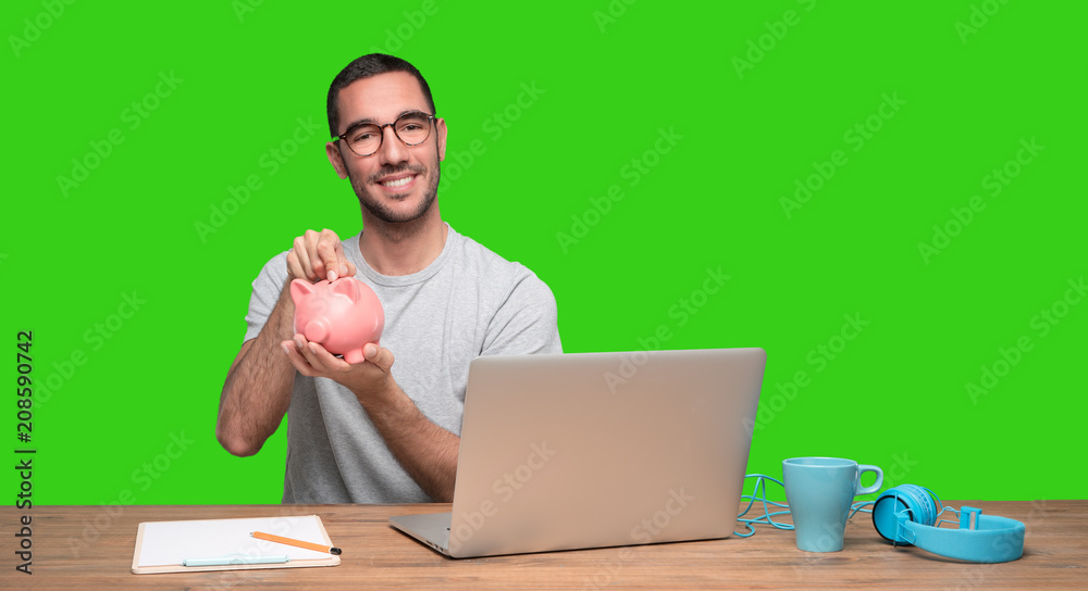 Happy young man sitting at his desk and holding a piggy bank - Green background Happy young man sitting at his desk and holding a piggy bank - Green background