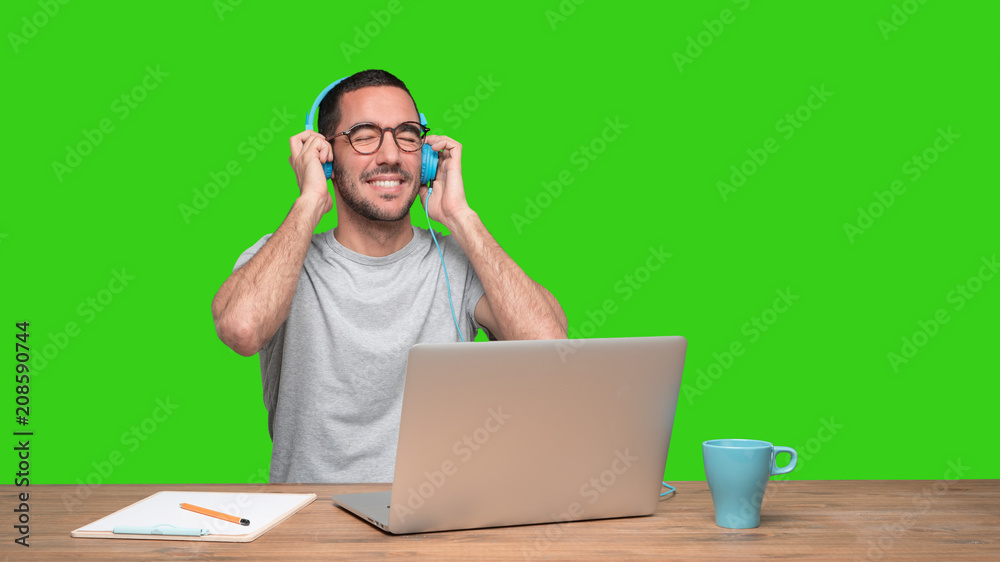 Happy young man sitting at his desk and using a headphones - Green background Happy young man sitting at his desk and using a headphones - Green background