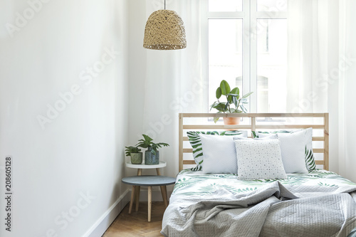 Stylish and modern sunny bedroom with plants , floral pattern bedding and pilows. Space with white walls and brown wooden parquet. 