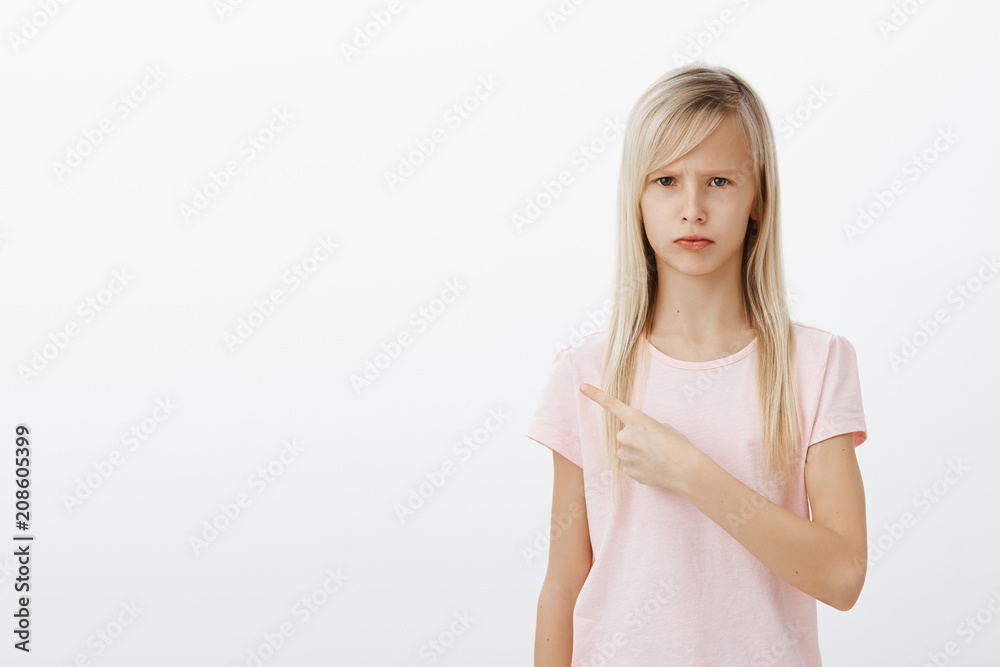 Studio shot of upset displeased adorable female child with blond hair, sulking and frowning while pointing at upper left corner, being upset and offended, standing over gray background moody Studio shot of upset displeased adorable female child with blond hair, sulking and frowning while pointing at upper left corner, being upset and offended, standing over gray background moody