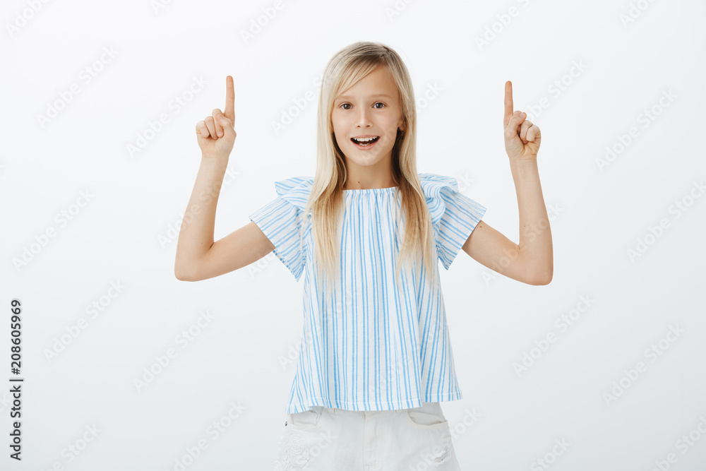 Studio portrait of happy good-looking young girl with blond hair, feeling cheerful while raising index fingers and pointing upwards, posing for advertisement or enjoying great sunny weather Studio portrait of happy good-looking young girl with blond hair, feeling cheerful while raising index fingers and pointing upwards, posing for advertisement or enjoying great sunny weather