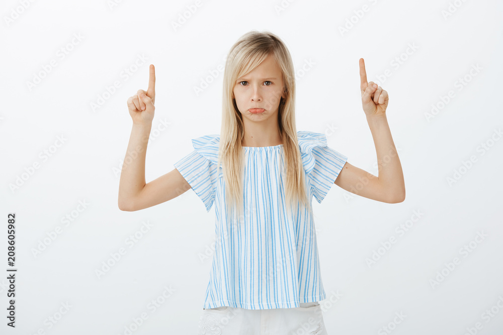 Portrait of offended angry caucasian female child with long blond hair, pouting and sulking, raising index fingers and pointing up, seeing something disappointing and insulting over gray wall