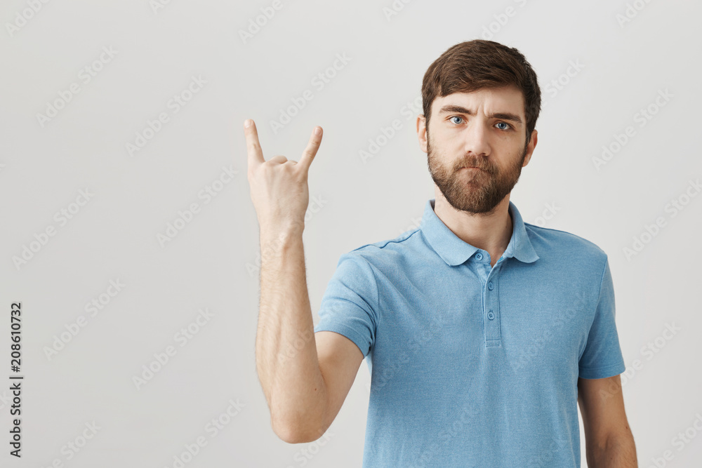 Rock n roll is alive. Portrait of serious cool european bearded man showing rock gesture with raised hand, expressing confidence and firm attitude to attend concert of favorite band Rock n roll is alive. Portrait of serious cool european bearded man showing rock gesture with raised hand, expressing confidence and firm attitude to attend concert of favorite band