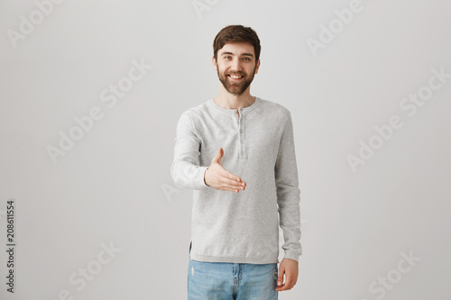 Give me firm handshake. Studio shot of emotive attractive man with beard pulling hands towards camera to greet business partner, smiling confidently while posing over gray background