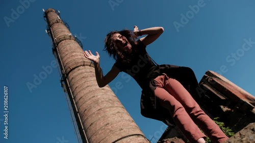A young woman is standing against the factory stack and joyfully disarranges her hair from the below view