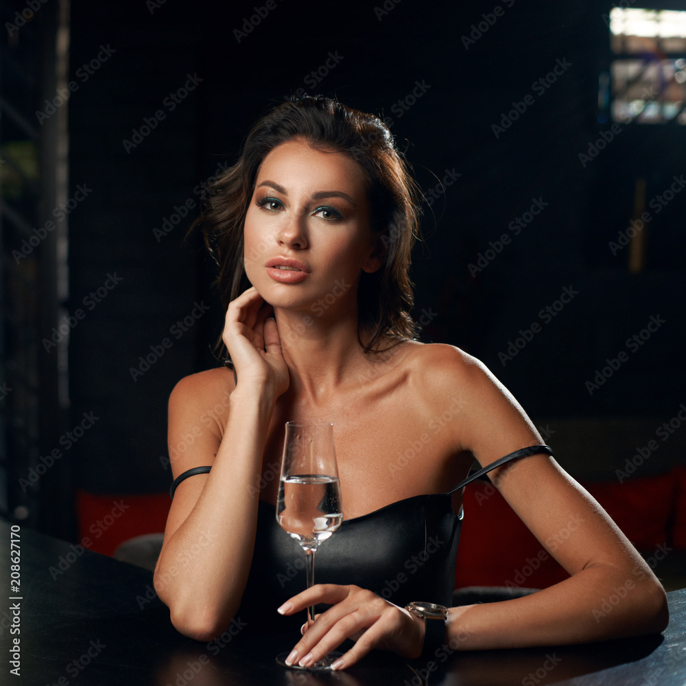 Elegant young woman sitting in bar and having good time alone. Pretty brunette girl in black leather dress. Lady drinking glass of wine.