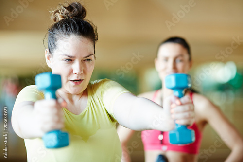 Young overweight female trying hard while exercising with dumbbells with coach on background