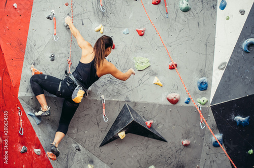 Fit sporty student girl equipped with safety rope and harness moving up at rock climbing wall at the gym. Red and grey colour background