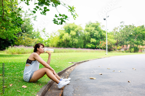 Beautiful asian woman is drinking electrolyte beverage or water during attractive beautiful woman is resting and sitting under a tree at public park. Charming beautiful girl lost sweat while exercise photo