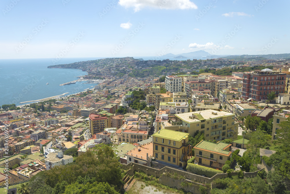 Fototapeta premium Beautiful aerial view from hill top fort Sant'Elmo on Naples gulf coast in Italy. East old town Naples landscape photo with blue sky, colorful buildings and island behind.