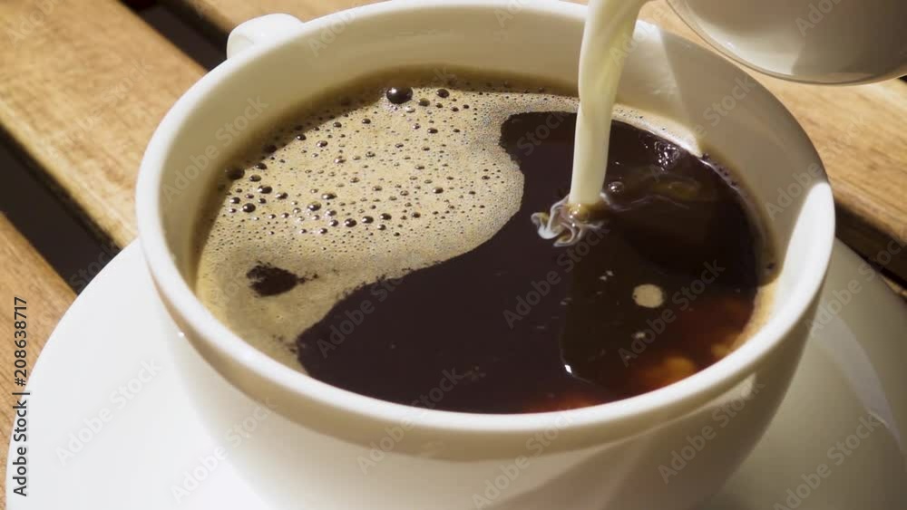Milk from a milkman pour into a porcelain cup and saucer with black coffee on a wooden table in the morning for breakfast slow motion Milk from a milkman pour into a porcelain cup and saucer with black coffee on a wooden table in the morning for breakfast slow motion
