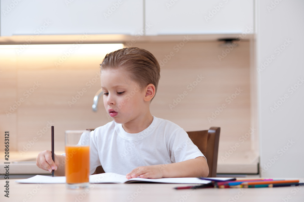 Little cute boy in white T-shirt paint the coloring book pencil at kitchen table next to glass of freshly squeezed juice Little cute boy in white T-shirt paint the coloring book pencil at kitchen table next to glass of freshly squeezed juice