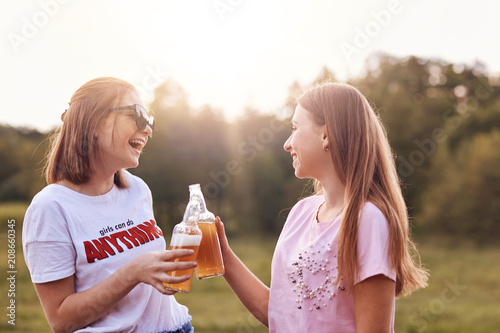 Best female friends toast with bottles of cold beer, have fun together, spend free time outdoor, spend summer holidays in countryside, laugh joyfully, enjoy relaxation. People and friendship concept